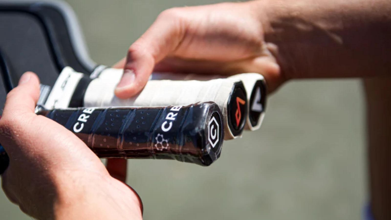 Hai tay đang cầm ba cây vợt pickleball với tay cầm được quấn băng màu đen và trắng, nổi bật với logo và các họa tiết trên đầu tay cầm.