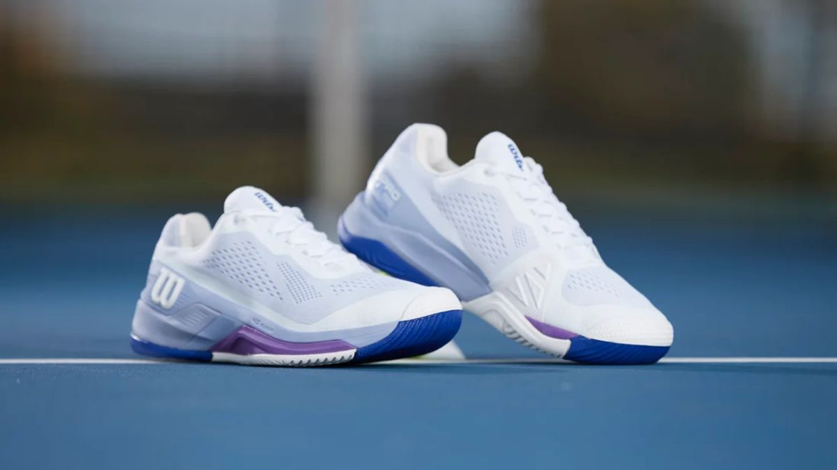 Một đôi giày tennis Wilson màu trắng với điểm nhấn màu tím và xanh ở đế, được đặt trên sân tennis màu xanh.