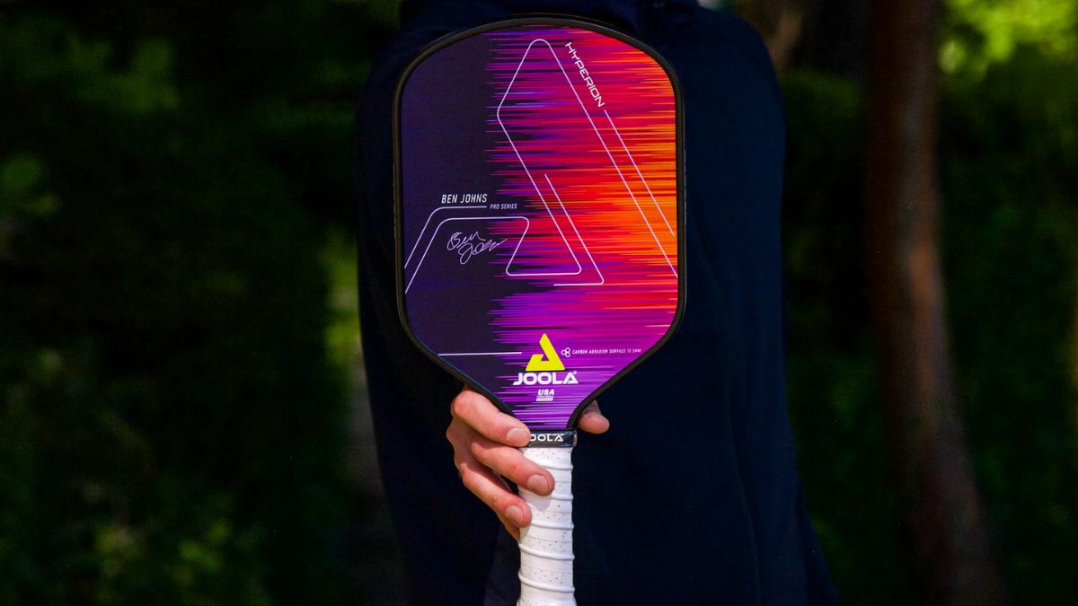 Một người đang cầm vợt pickleball JOOLA Hyperion Ben Johns Pro Series, với thiết kế màu sắc nổi bật gồm tông tím, đỏ và cam, tay cầm màu trắng, đứng trước khung cảnh thiên nhiên mờ phía sau.