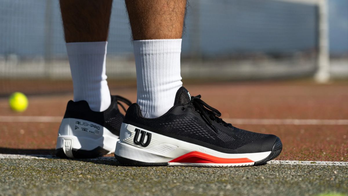 Giày tennis Wilson màu đen với chi tiết trắng và đỏ, được mang bởi một người kết hợp với tất trắng, trên sân tennis ngoài trời.