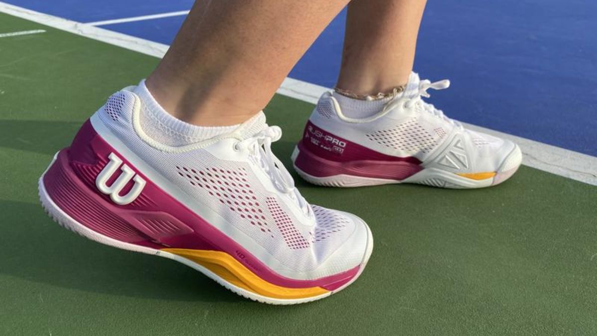 Giày tennis Wilson màu trắng với điểm nhấn màu hồng và vàng, được mang bởi một người trên sân tennis ngoài trời.
