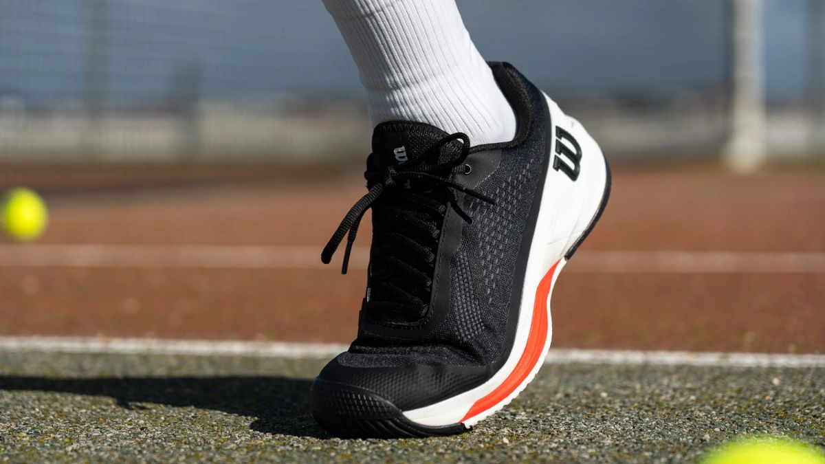 Giày tennis Wilson màu đen với chi tiết trắng và đỏ, được mang bởi một người kết hợp với tất trắng, trên sân tennis với bóng tennis màu vàng ở phía xa.