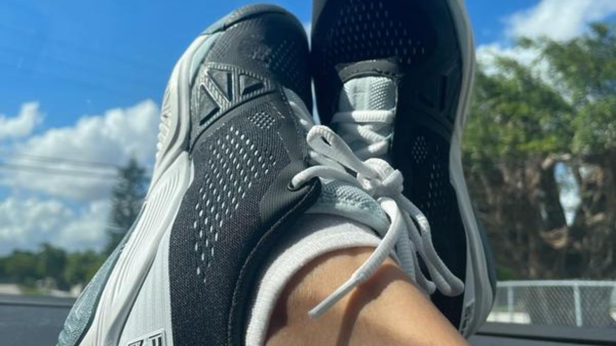 Một đôi giày tennis Wilson Rush Pro 4.0 màu đen với chi tiết xám, được mang bởi một người đang thư giãn trên ghế xe hơi, dưới bầu trời xanh trong.