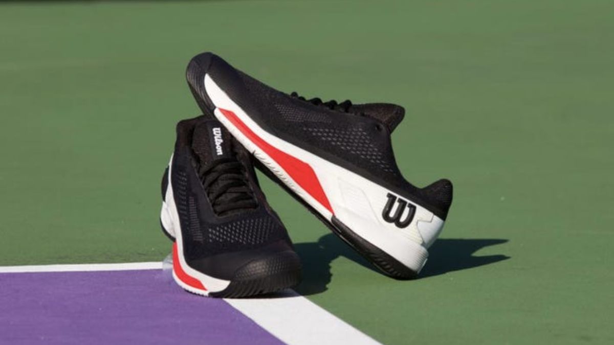 Một đôi giày tennis Wilson màu đen với chi tiết đỏ và trắng, được đặt trên sân tennis có nền xanh và tím.