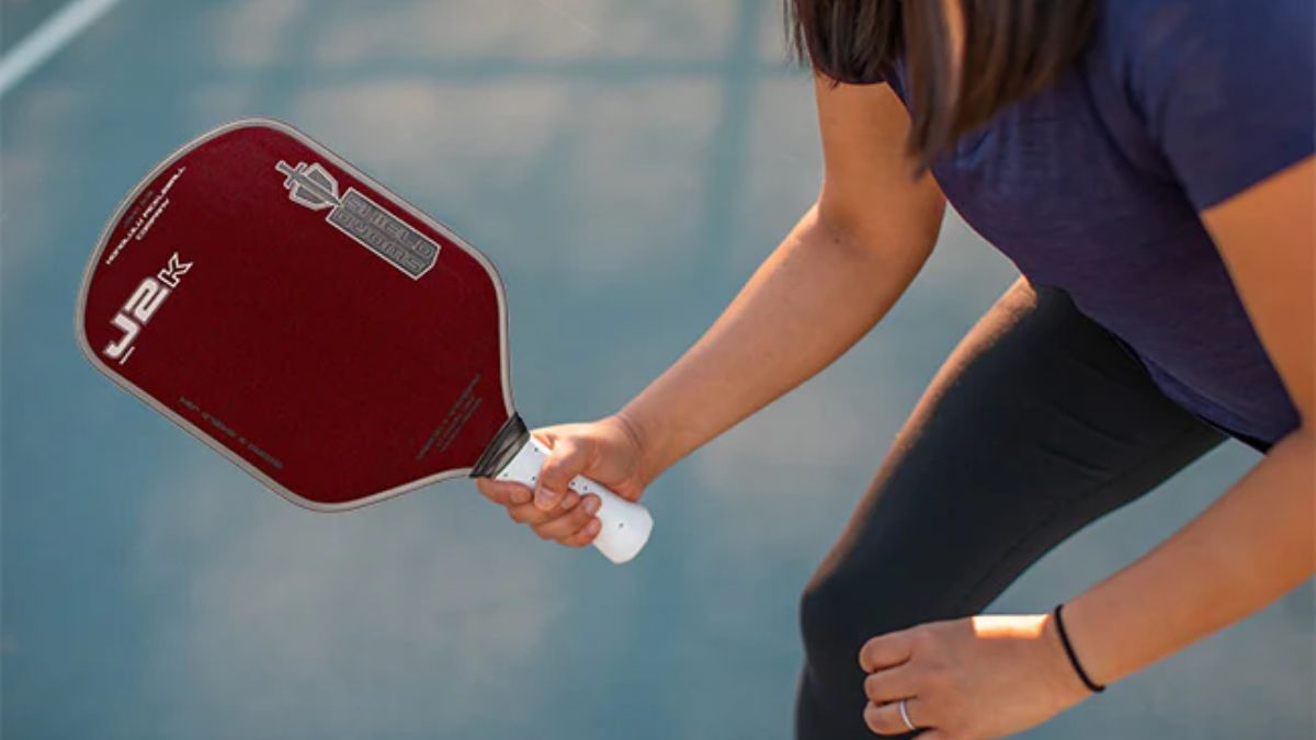 Một người chơi pickleball đang cầm vợt Honolulu Pickleball J2K màu đỏ sẫm với tay cầm màu trắng, chuẩn bị đánh trên sân ngoài trời dưới ánh sáng tự nhiên.