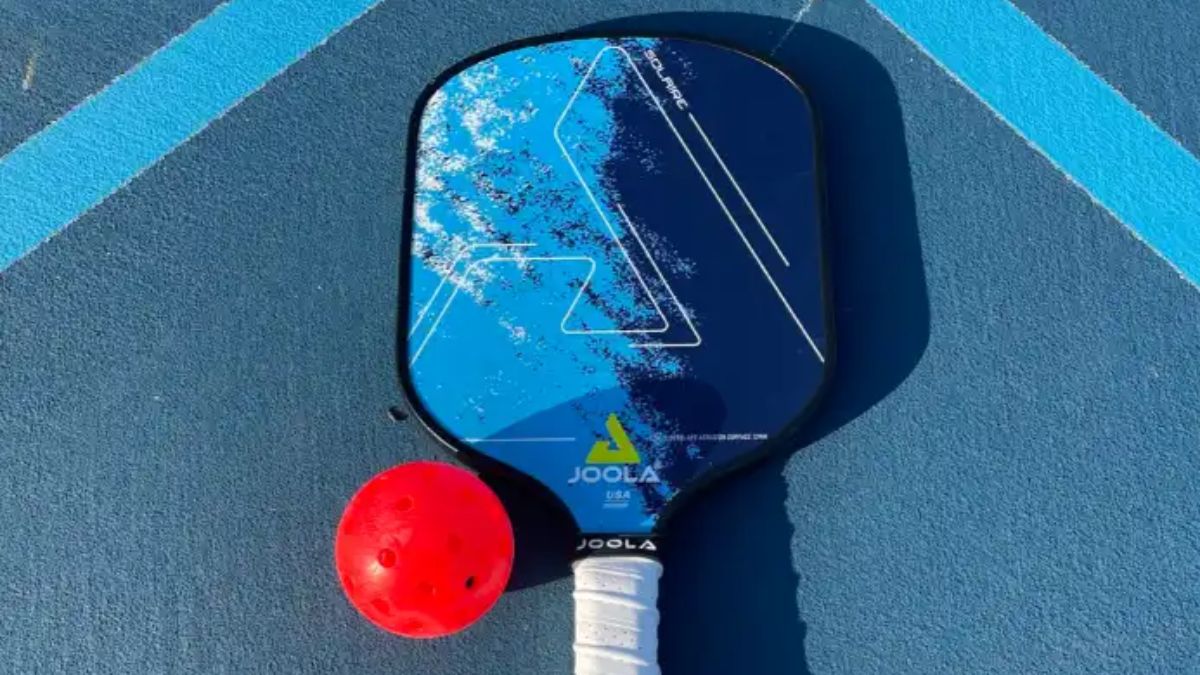 Vợt pickleball Joola Solaire FAS 13 với thiết kế màu xanh và họa tiết độc đáo, được đặt trên sân xanh cùng một quả bóng pickleball màu đỏ nổi bật.