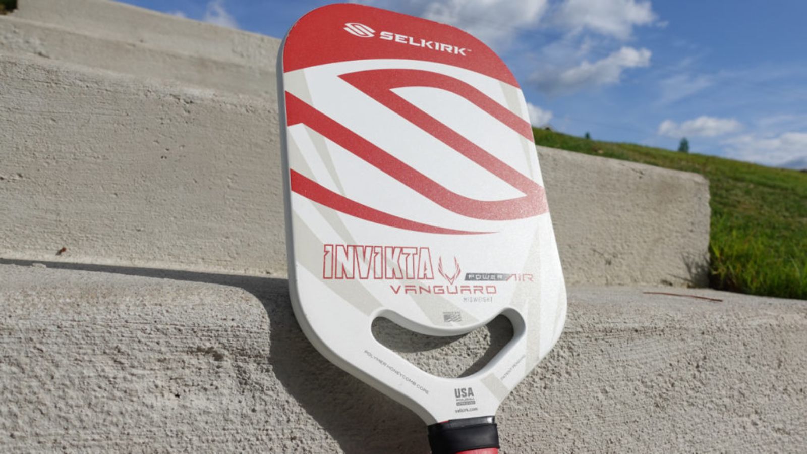 Vợt pickleball Selkirk Vanguard Power Air Inviktá, thiết kế màu đỏ và trắng, được đặt trên bậc thang bê tông với khung cảnh bầu trời trong xanh và cỏ xanh phía sau.