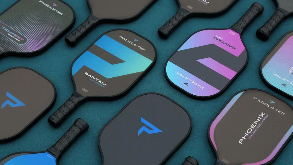 Một bộ sưu tập vợt pickleball được bày trên bề mặt màu xanh lam, với nhiều thiết kế và màu sắc khác nhau, bao gồm các mẫu Paddletek Bantam và Phoenix.